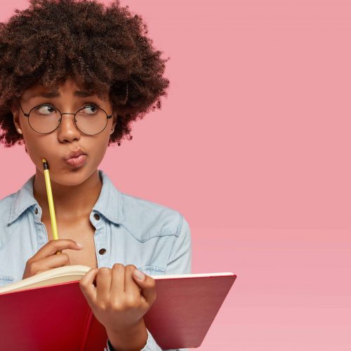 Pensive confused dark skinned woman purses lips in bewilderment, writes notes in notepad, thinks on creating new article for personal blog, wears round transparent glasses poses indoor with copy space