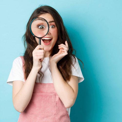 Happy young woman looking through magnifying glass with excited face, found or search something, standing over blue background.